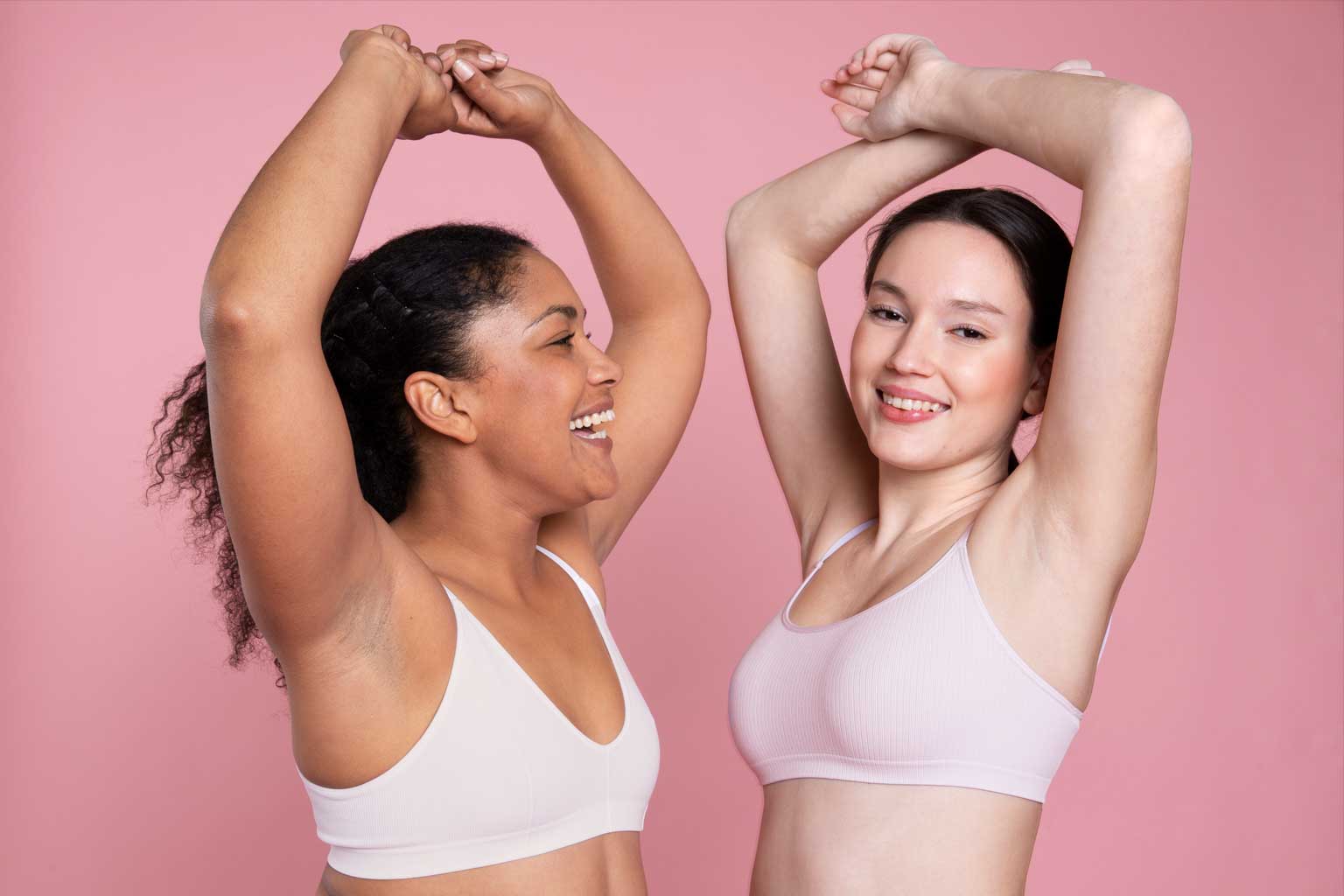 Two women displaying smooth underarms after laser hair removal treatment against a pink studio background.